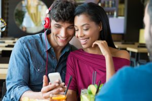 Closeup shot of young couple listening to music with mobile phone at coffee bar. Young man and african woman looking at smartphone at caf??. Happy young couple listening music with headphones.